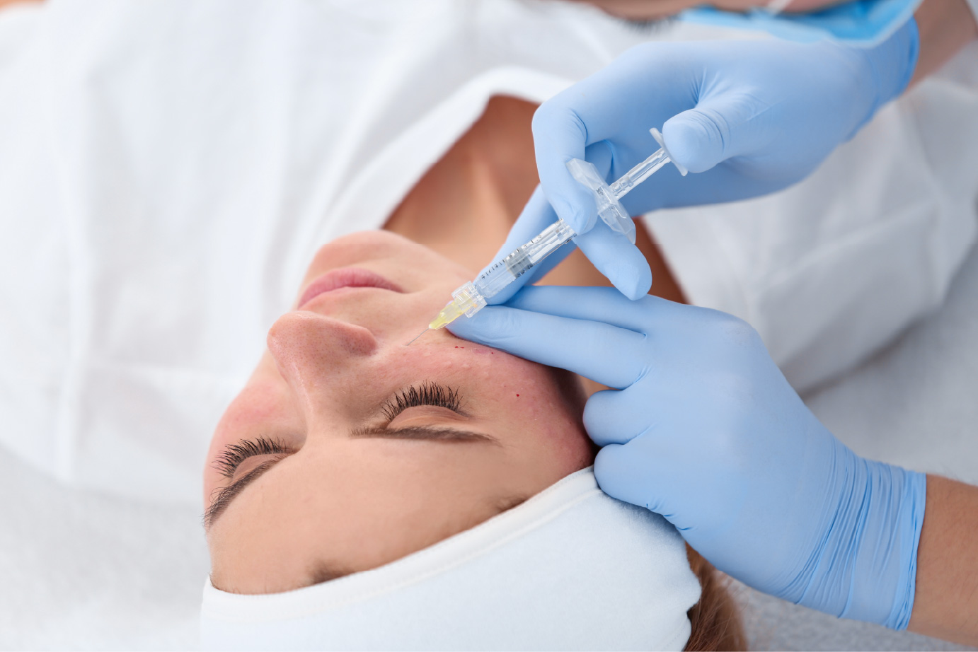 A person lying down with eyes closed while gloved hands hold a syringe near the cheek.